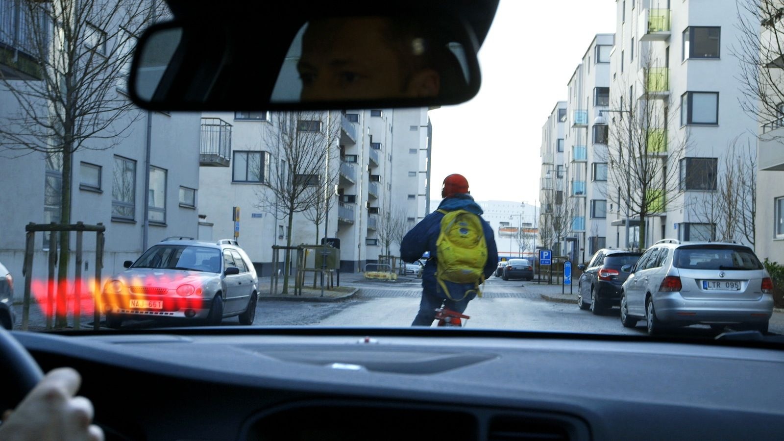 Driver's POV from inside a car: cyclist with a yellow backpack crossing ahead on an urban street, parked cars on both sides, silver hatchback braking ahead-left